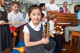 A young student in school uniform holds a jingle stick as her classmates play other percussive instruments behind her.