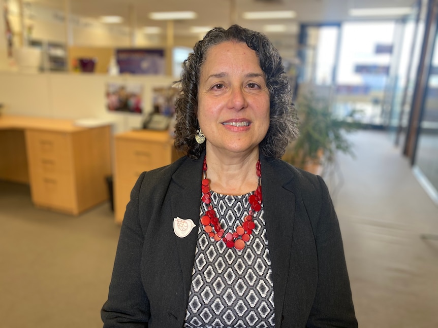 A smiling woman stands in her office, wearing a red necklace, gray with diamonds, a black and white top and a dark jacket.