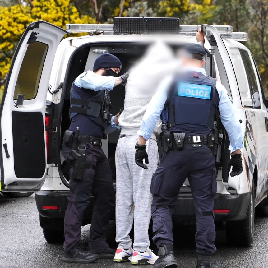 Police putting a man wearing a grey tracksuit into a paddy wagon pictured from behind.