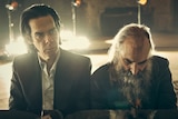 Nick Cave and Warren Ellis, two middle-aged men, sit at a piano together, Cave looking into the distance, Ellis at the keys