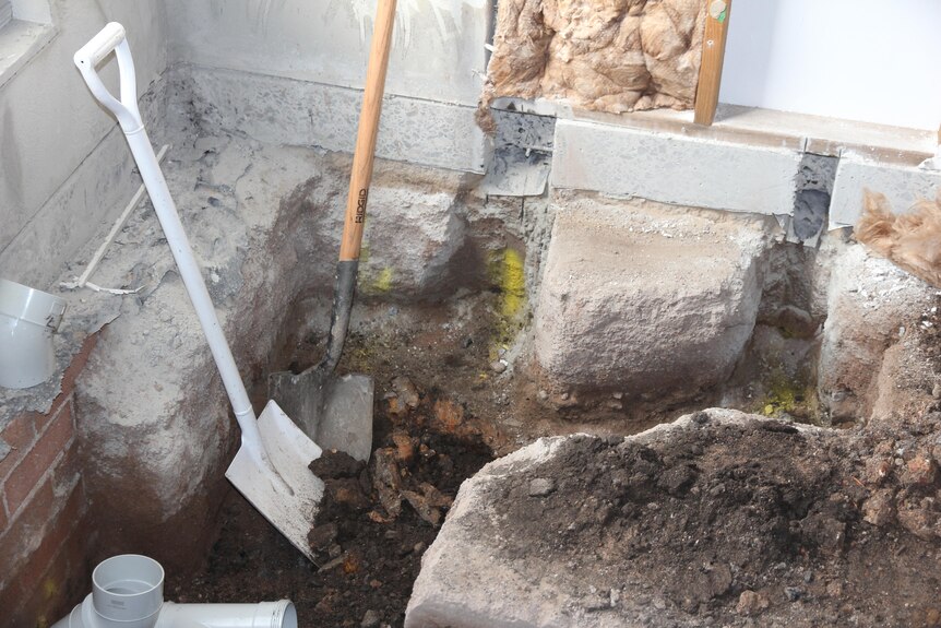 Two shovels lean up against wall, dirt hole in ground, bits of concrete next to holes.