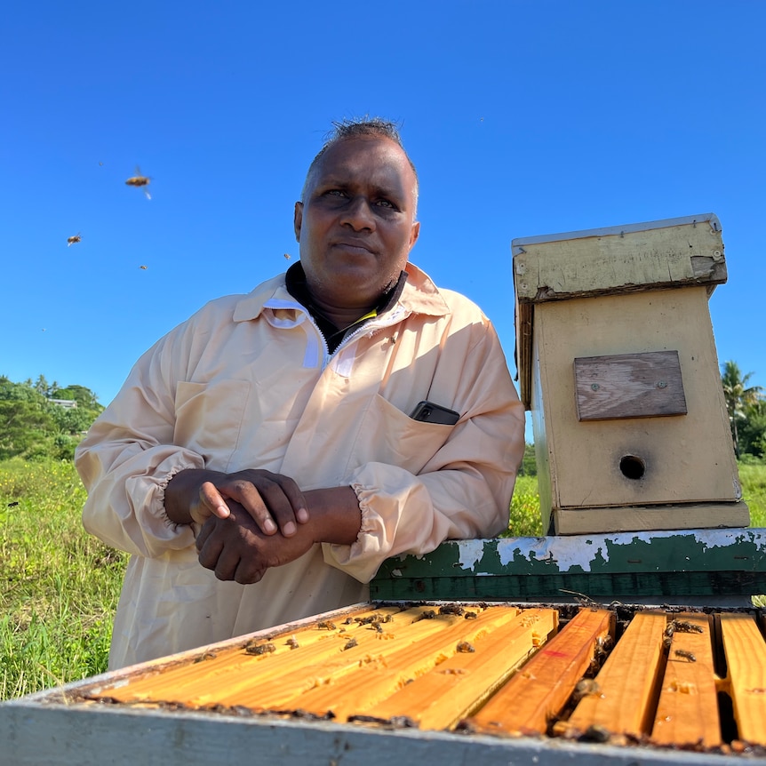 Fiji beekeeper in beekeeping suit leans on hive. 