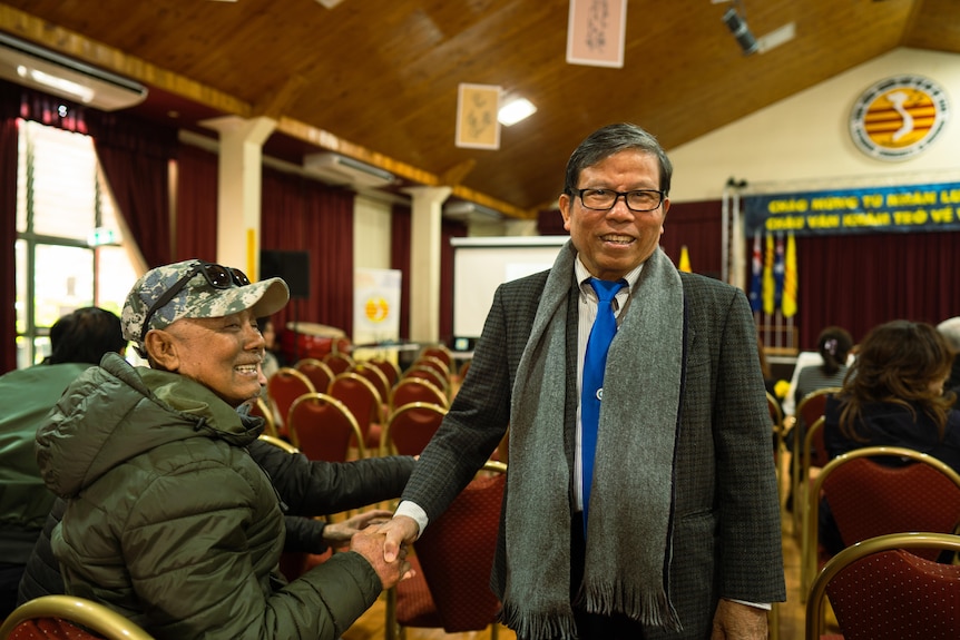 Asian man wearing a suit with a blue tie and scarf shakes hands with another man. They're in a community hall.