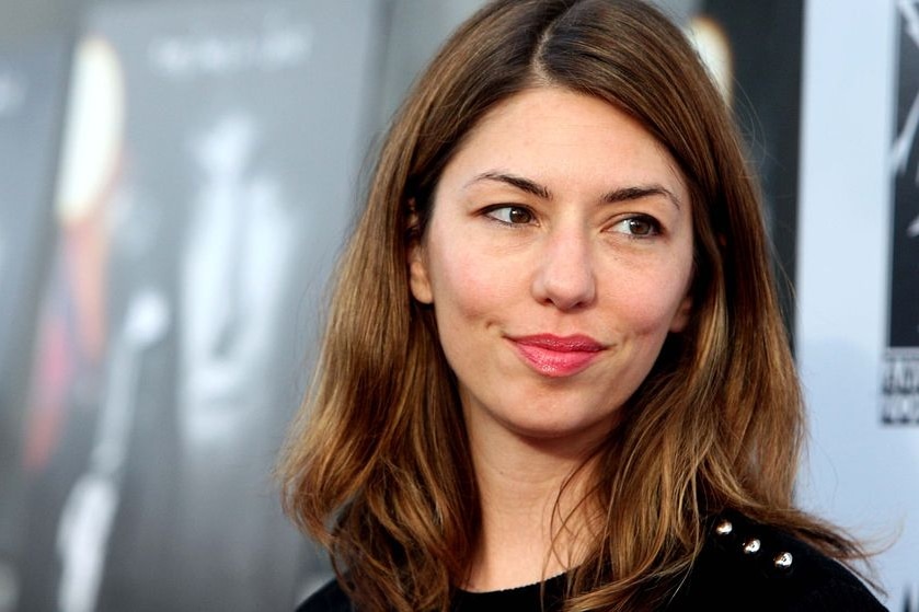 Sofia Coppola, a white middle-aged woman with dark brown hair is photographer on a red carpet, smiling at someone off-camera.