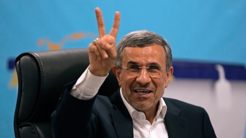 An older Iranian man with grey hair in a suit holds up two fingers and smiles in front of a blue background.