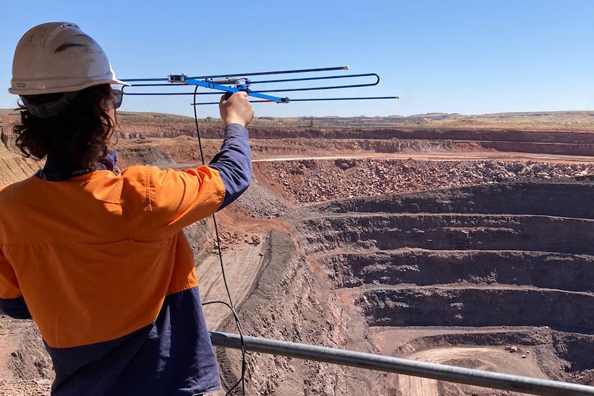 A man in a high-vis shirt and hard hat holds up an antenna towards a large open cut Manganese mine in the Pilbara.