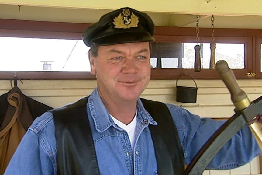 A fair-skinned, middle-aged man in denim shirt and ship captain hat holds the steering wheel of a paddle steamer.