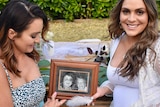 Two women sit together holding a photo of their mother and grandmother. 