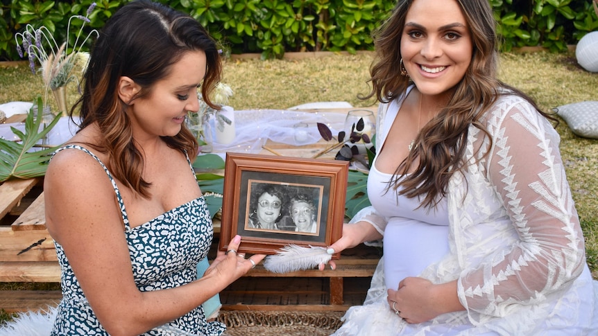 Two women sit together holding a photo of their mother and grandmother. 