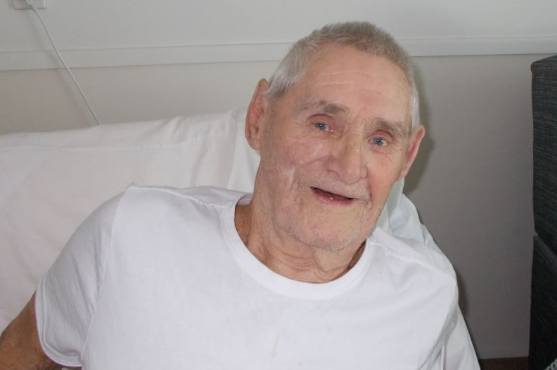 An elderly man wearing a white tshirt sits in a nursing home bed and smiles at the camera