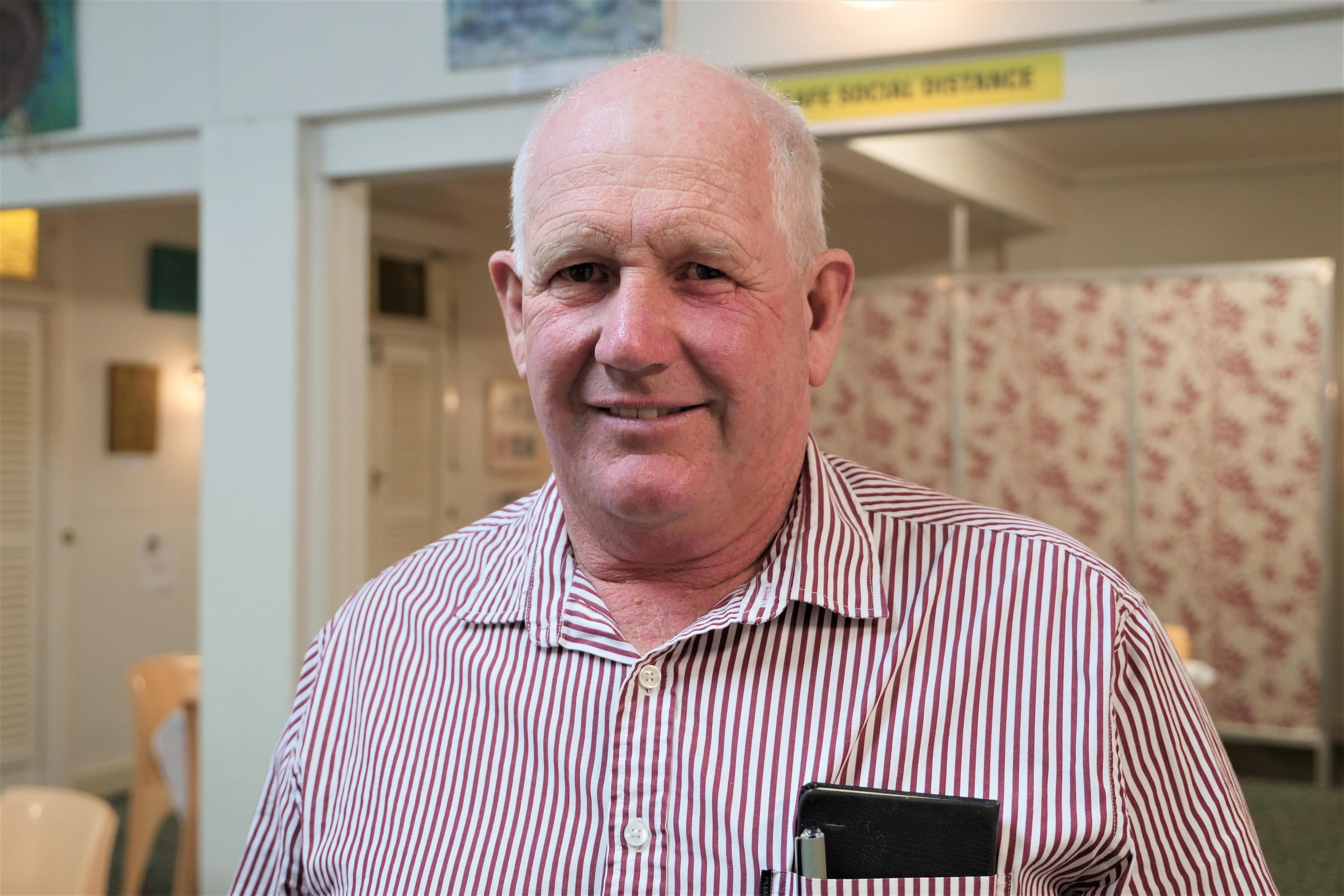 An older man has a small smile. He's standing in front of a wall with old school red and white wallpaper.