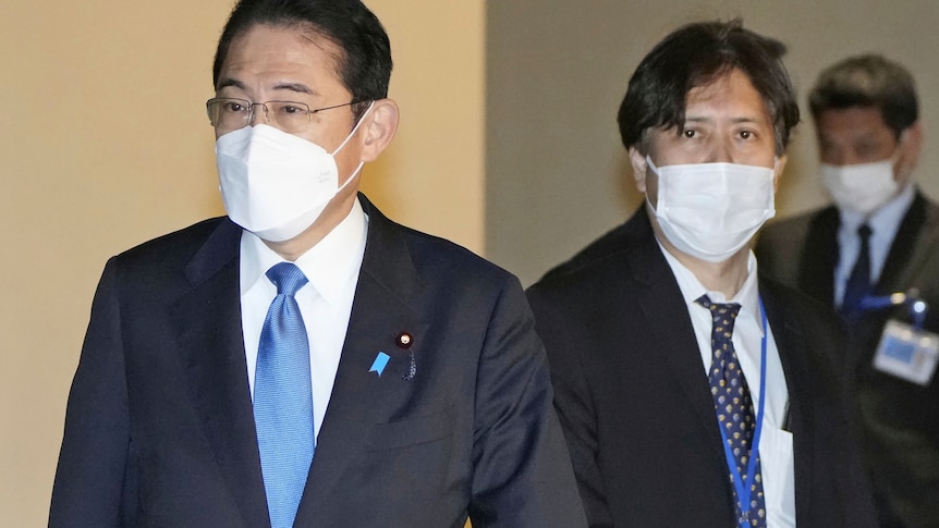 Two Japanese men in suits wearing face masks walk out of room. 