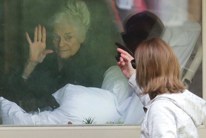 An elderly  woman in a hospital bed waves through a window to her daughter outside.