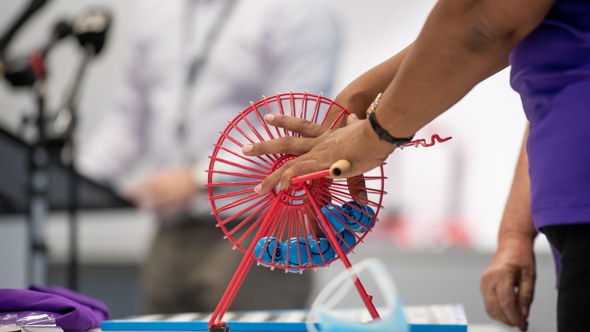 A person's hands picking a numbered blue ball out of a red plastic bingo barrel.