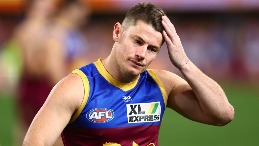 A Brisbane Lions AFL player puts his left hand to his head.
