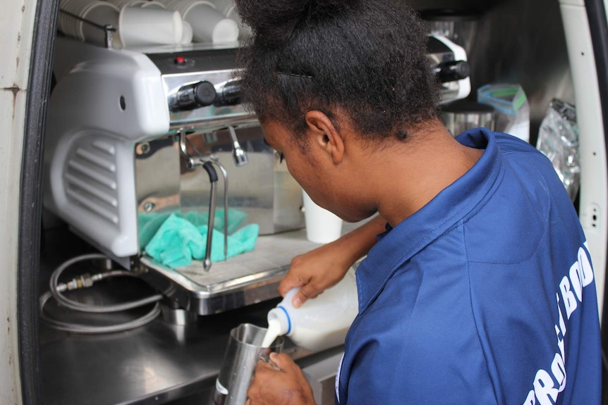 A young women stands at a coffee making machine pouring milk into a jug