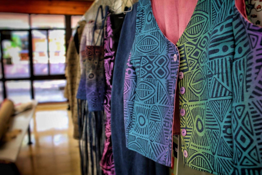 Bright and colourful garments with Tiwi Islands prints hang on a wall.