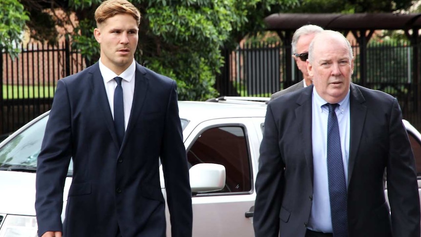 A young man in a suit, flanked by two older, similarly attired men, walks in front of a 4WD parked near a school.