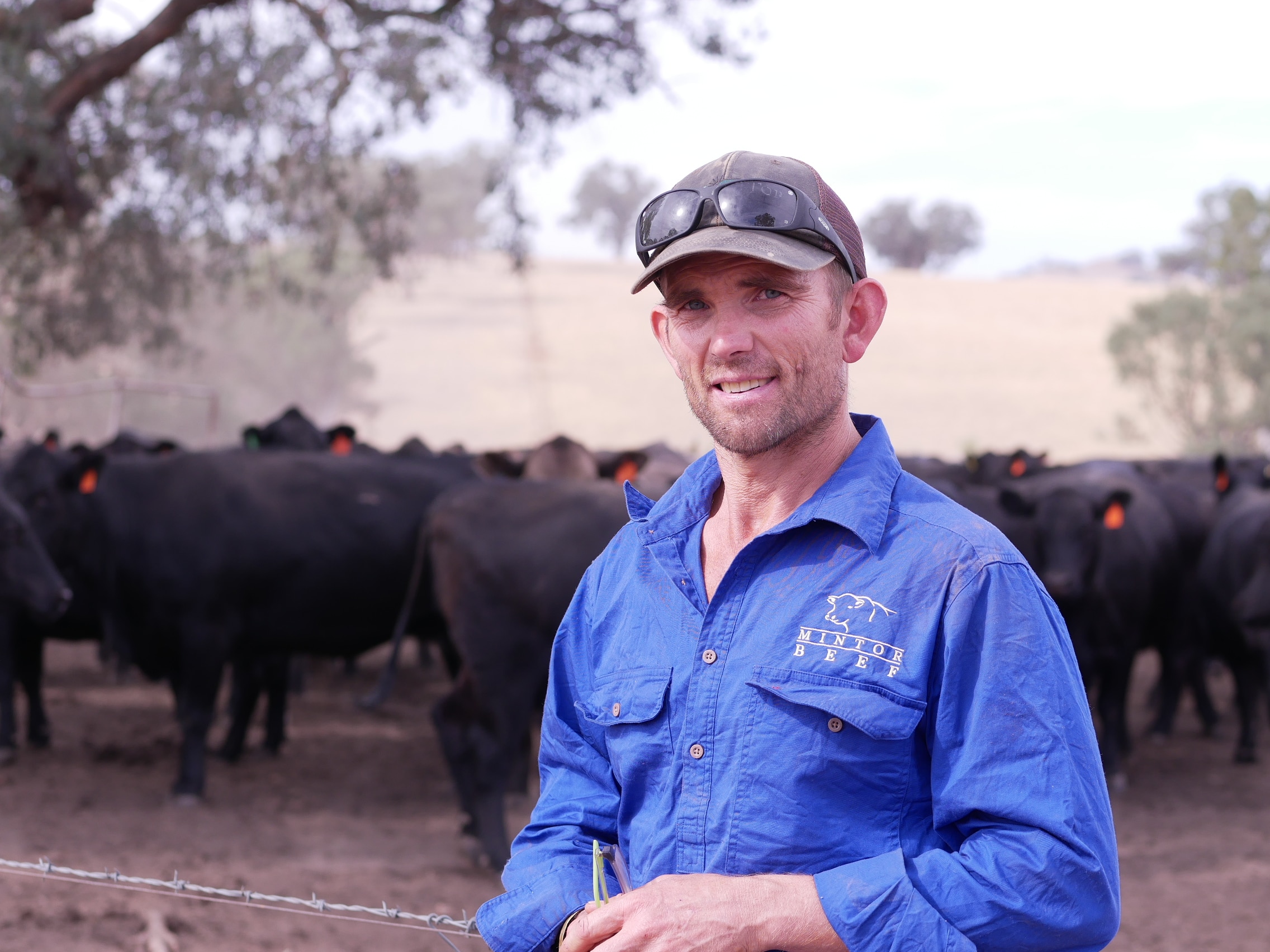 Un hombre con una camisa azul sonríe a la cámara, se pueden ver vacas Angus al fondo