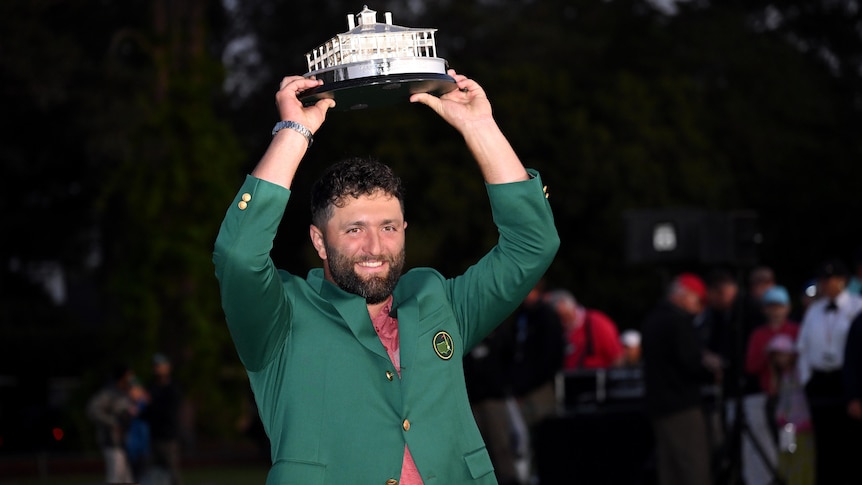 Jon Rahm smiles while holding the Masters trophy over his head, wearing the green jacket