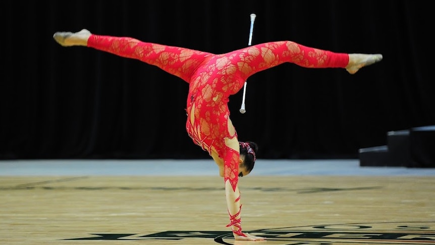 A girl in a red costume stands on one hand with her legs in the air in the splits