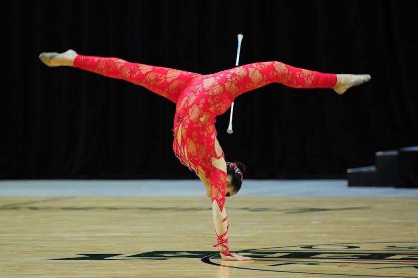 A girl in a red costume stands on one hand with her legs in the air in the splits