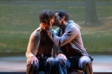 Two young white man wearing streetwear, sit on the lip of a stage and kiss in an embrace.