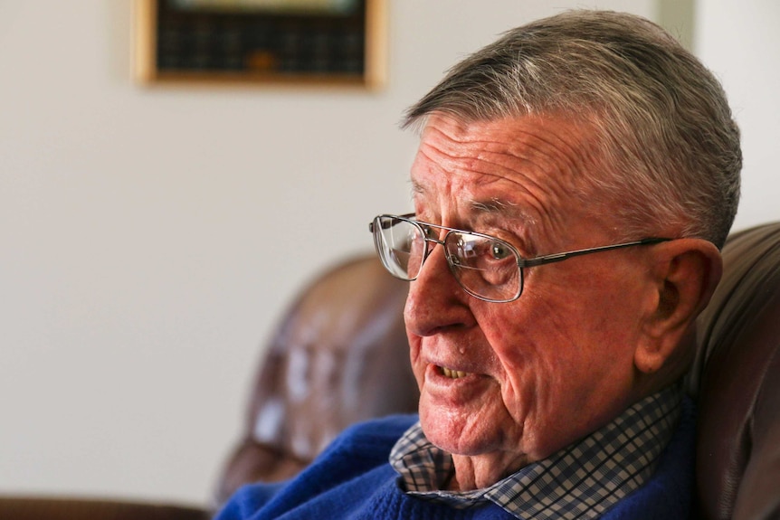 Older man with grey hair stares through his glasses, while sitting on a brown couch.