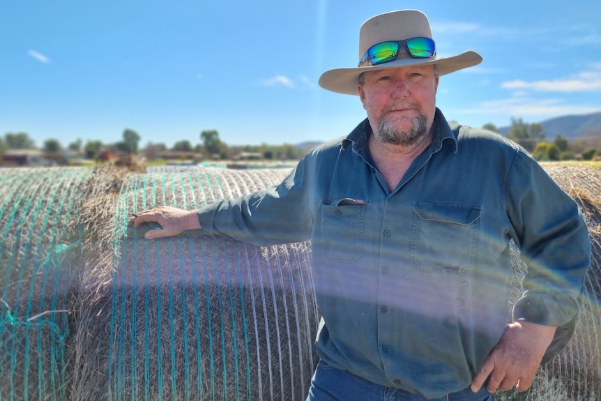 A man wearing a wide brimmed hat leans on a round hay bale and looks into camera.