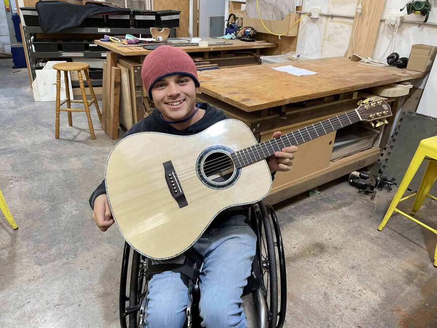 A smiling young man in a wheelchair shows off a guitar he made.