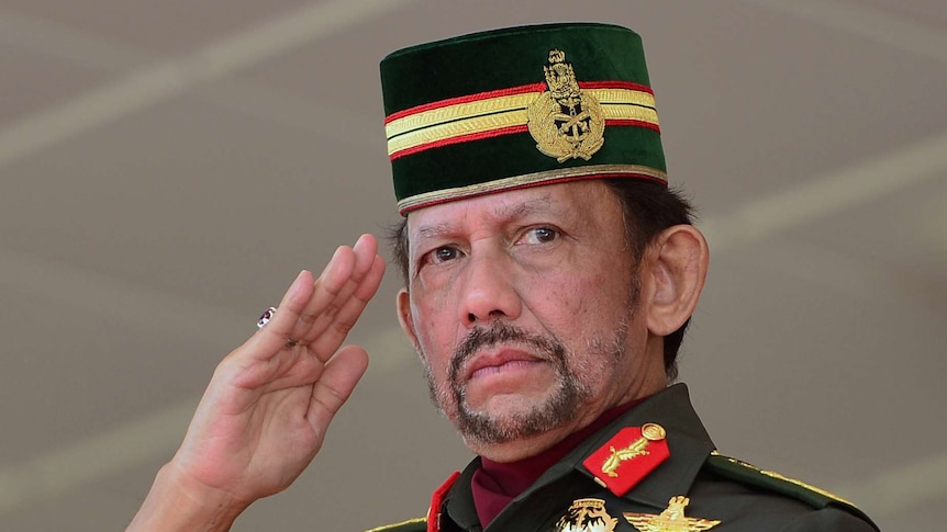 Sultan Hassanal Bolkiah salutes, wearing a decorated military uniform and cap. He has a large red ring on his right hand.