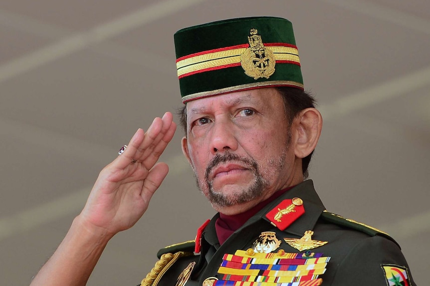 Sultan Hassanal Bolkiah salutes, wearing a decorated military uniform and cap. He has a large red ring on his right hand.