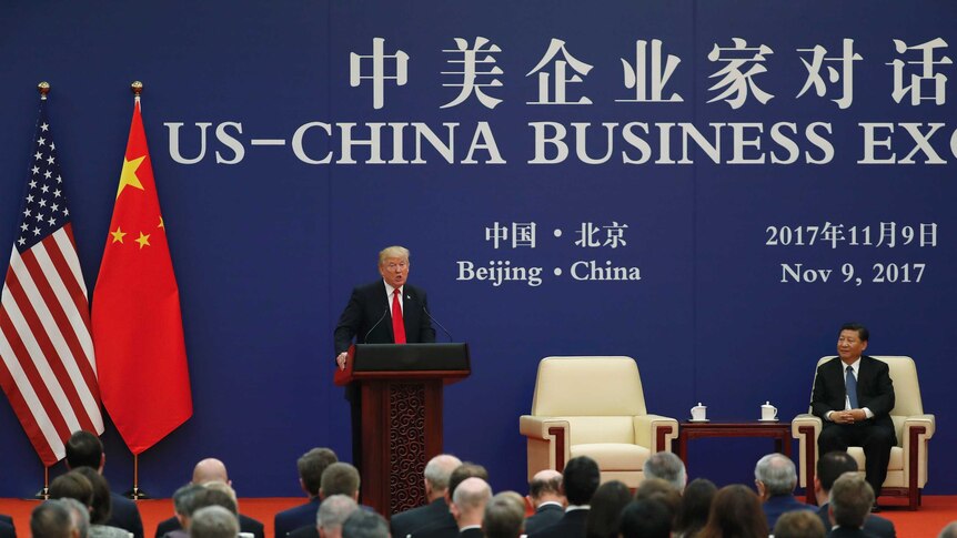 Donald Trump speaks at a podium while Xi Jinping sits down on his right.