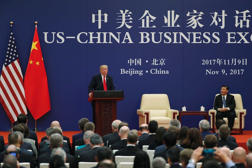 Donald Trump speaks at a podium while Xi Jinping sits down on his right.