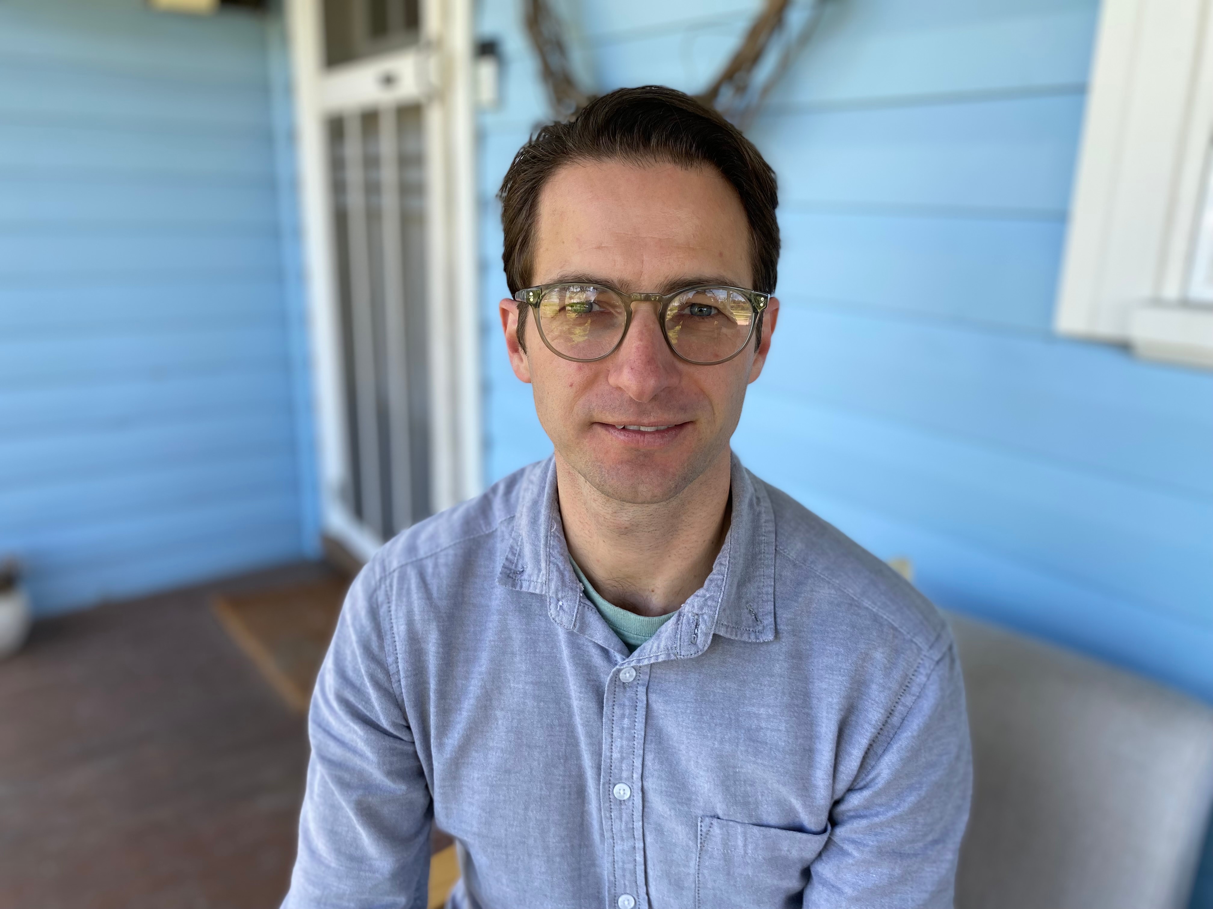 A man with glasses sits on his porch.