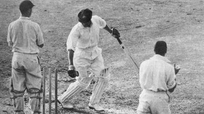Bradman looking back at his stumps with the bails off with the wicket keeper and slip behind him.