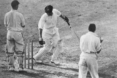 Bradman looking back at his stumps with the bails off with the wicket keeper and slip behind him.