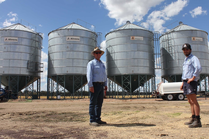 Two men smile at the camera with four large, silver silos in the background against a bright blue sky