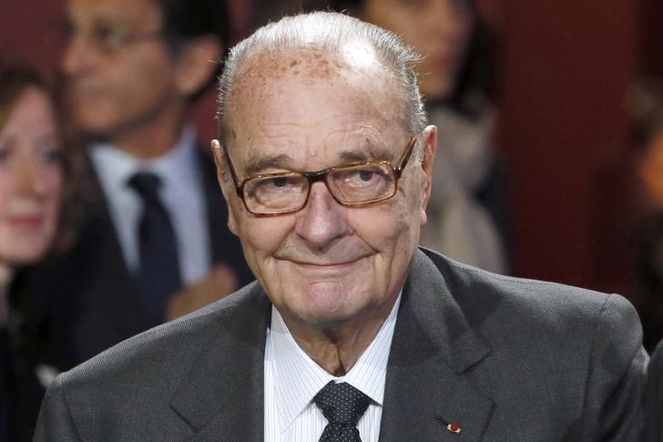 Jacques Chirac smiles as he looks to the right. He wears a dark suit with a dark tie and striped shirt and has glasses on.