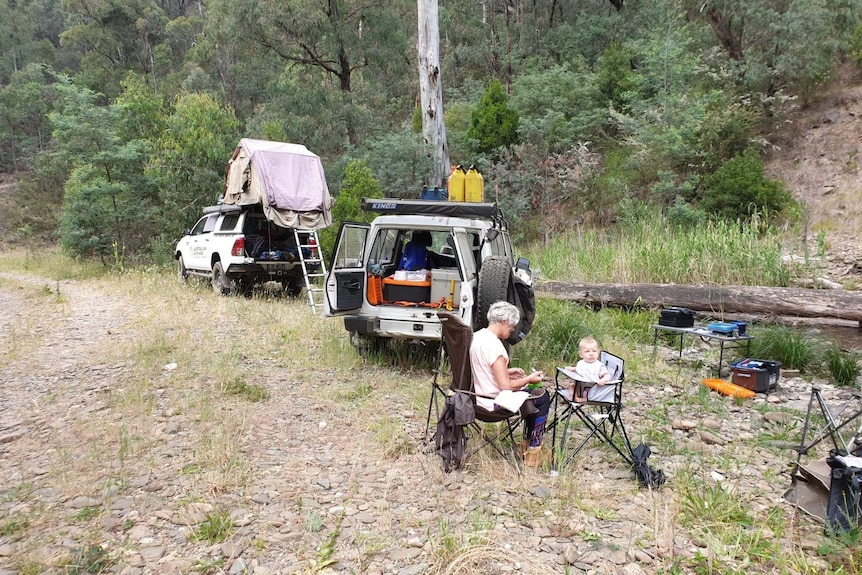 A grandmother and toddler sit in camping chairs surrounded by bush, with two 4WDs next door.