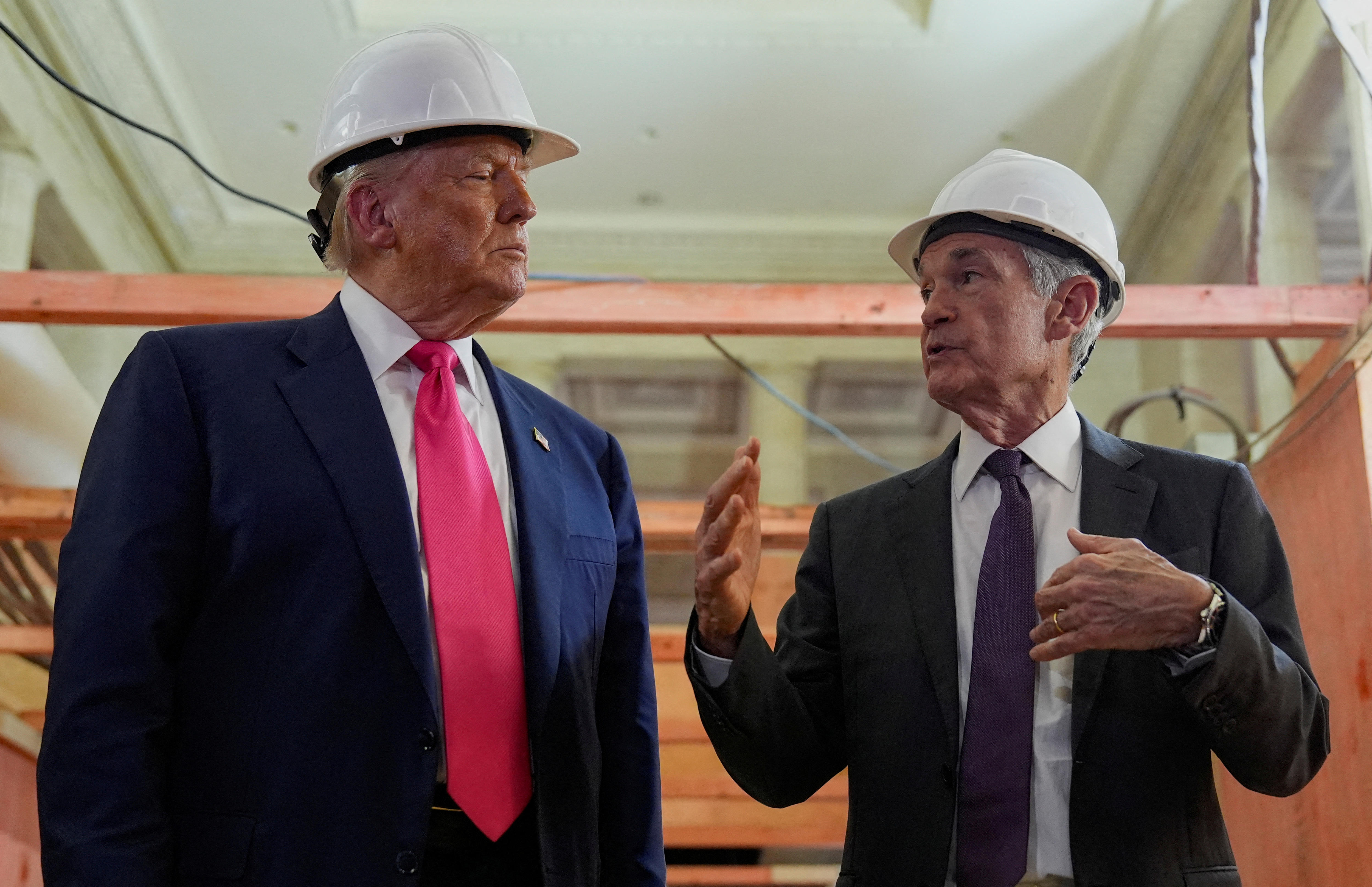 Donald Trump mira fijamente a un hombre más pequeño que habla con él mientras ambos usan cascos y trajes en un sitio de construcción.