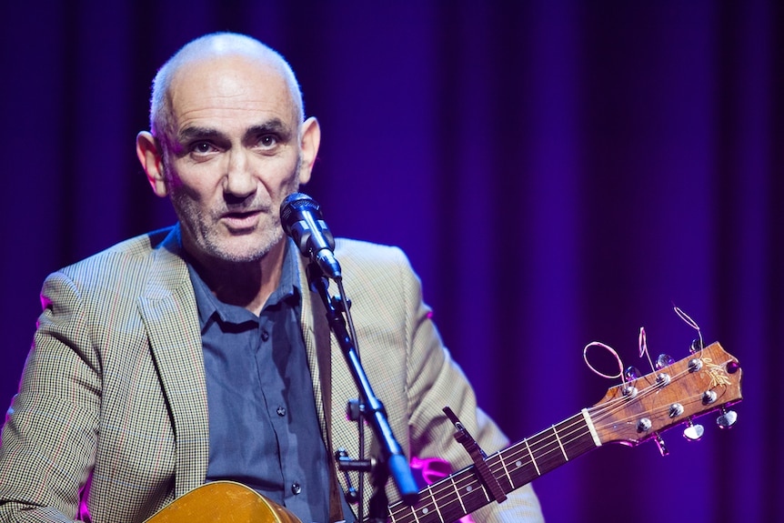 Paul Kelly on stage with an acoustic guitar, wears a light jacket, blue shirt, purple curtains behind.