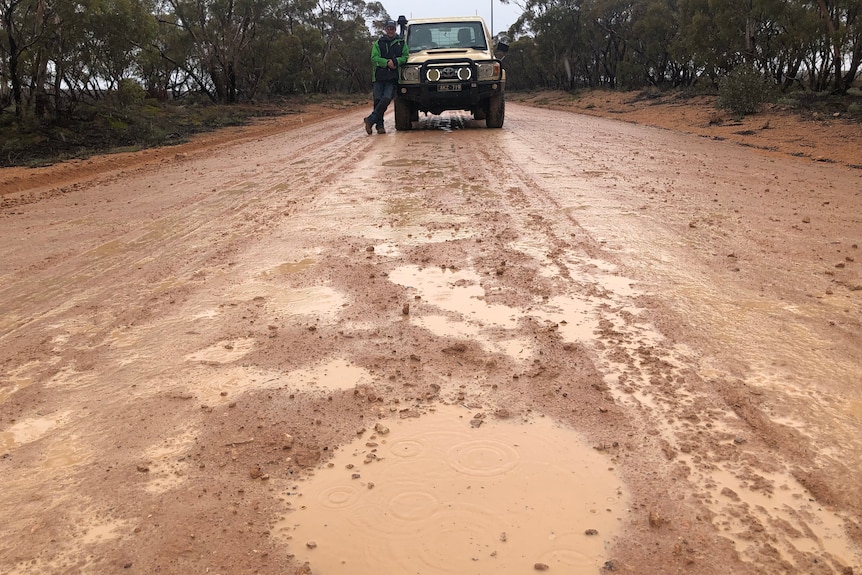 A man leans on a four wheel drive on a wet dirt road in front of a pot hole filled with water