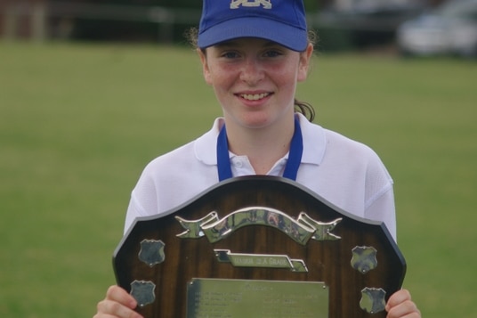 A young girl in cricket whites and a blue cap smiles while holding a shield. She has a medal around her neck