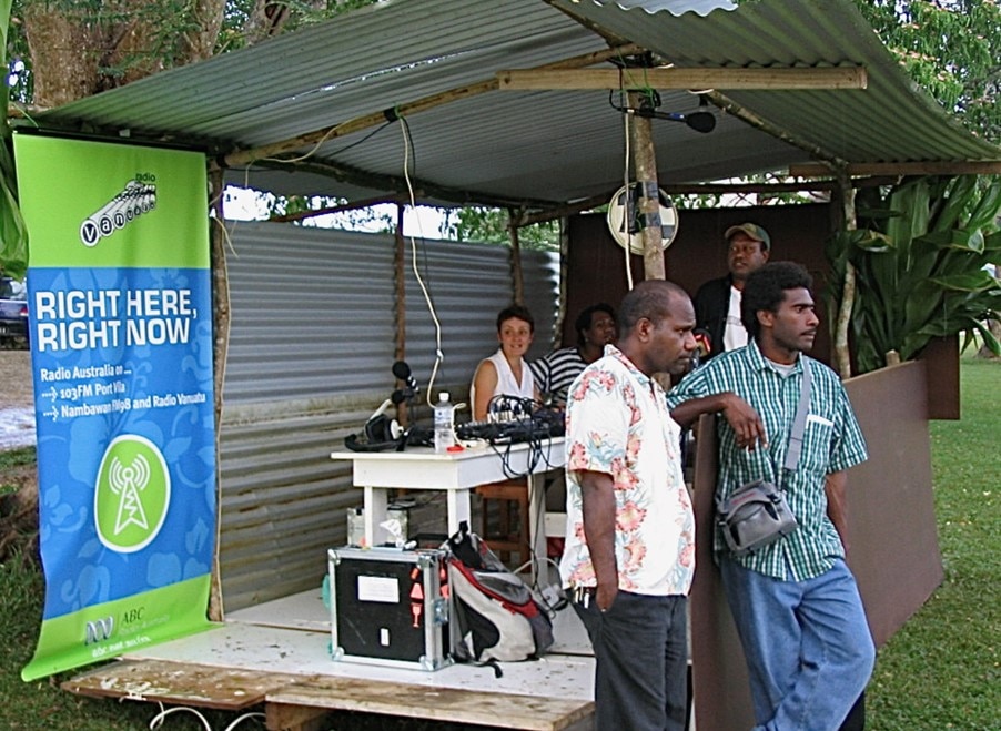 Woman broadcasting from an open tin shed with audio equipment set up on a table.