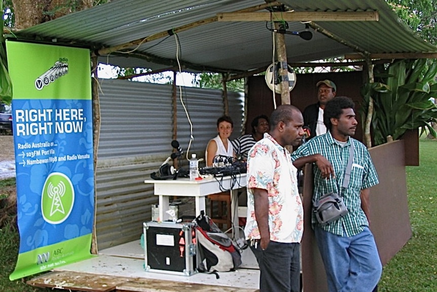 Woman broadcasting from an open tin shed with audio equipment set up on a table.