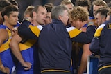 Adam Simpson speaks to his Eagles players in a tight huddle during a night AFL match, with his arms around two of them.