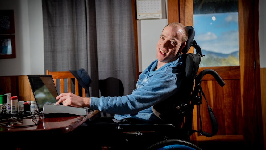 Nathan using a laptop in a homely-looking dining room, and smiling widely at the camera.