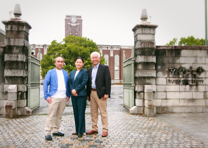 Three Japanese people in suits stand in front of a stone gate. 