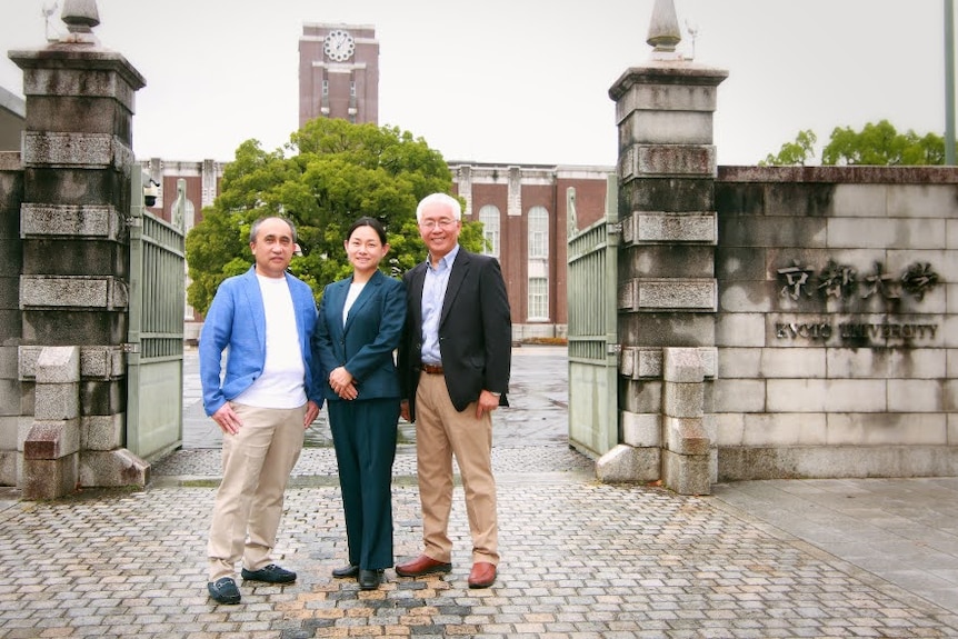 Three Japanese people in suits stand in front of a stone gate. 